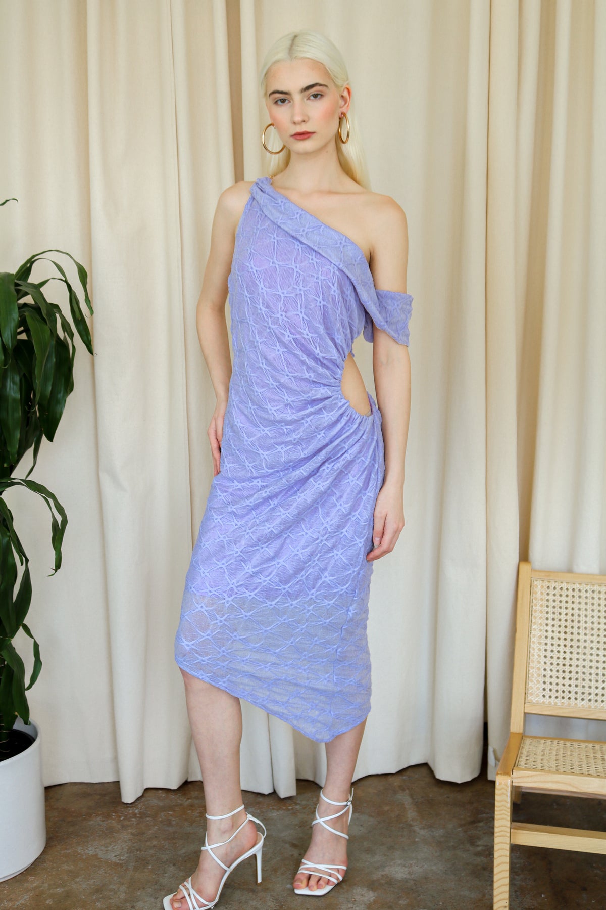 Model wearing a purple one-shoulder textured midi dress, standing beside a plant and a white cushioned chair.
