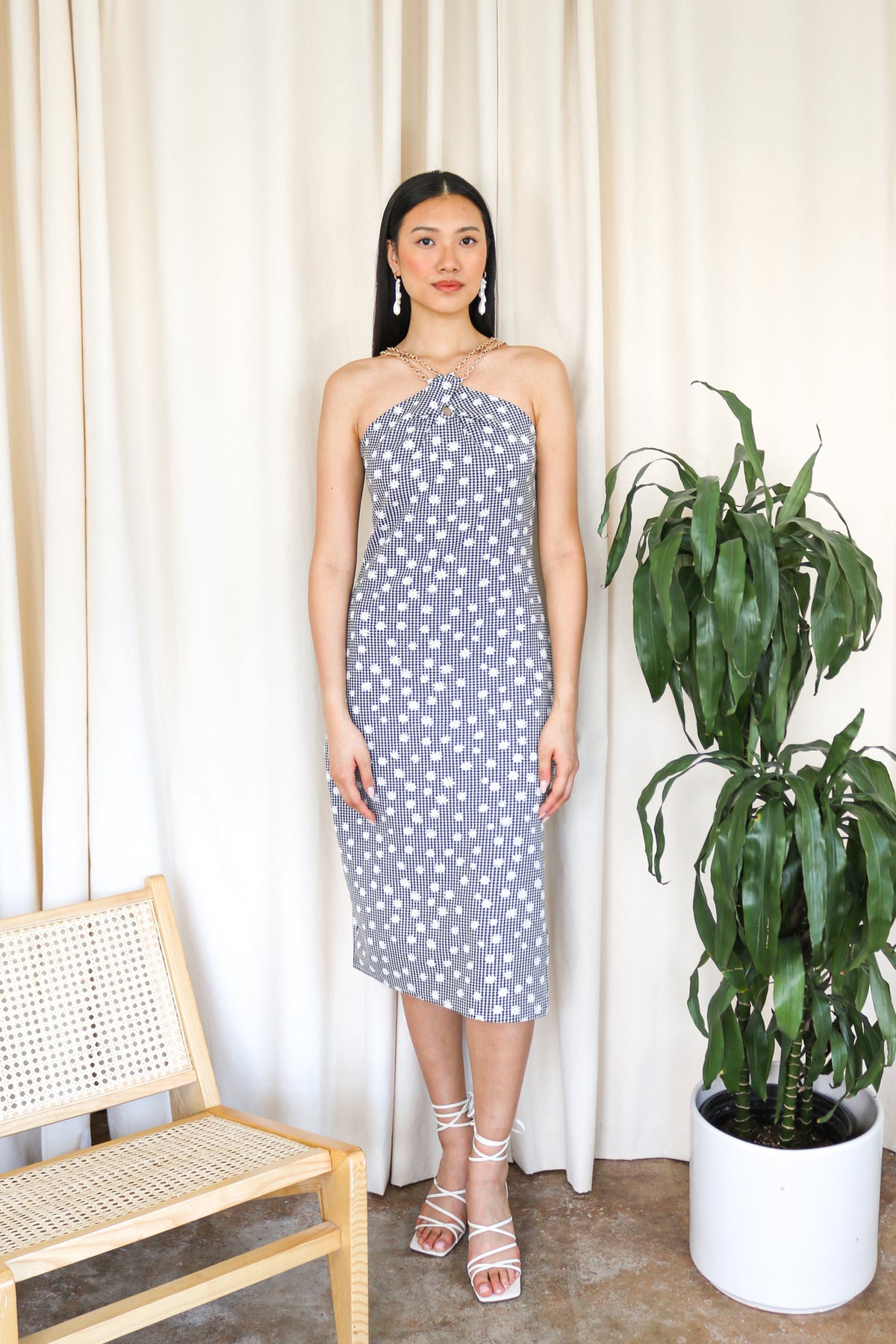 Model is wearing a black and white gingham midi dress with floral detail and criss cross gold chain detail around the neckline. Front facing.