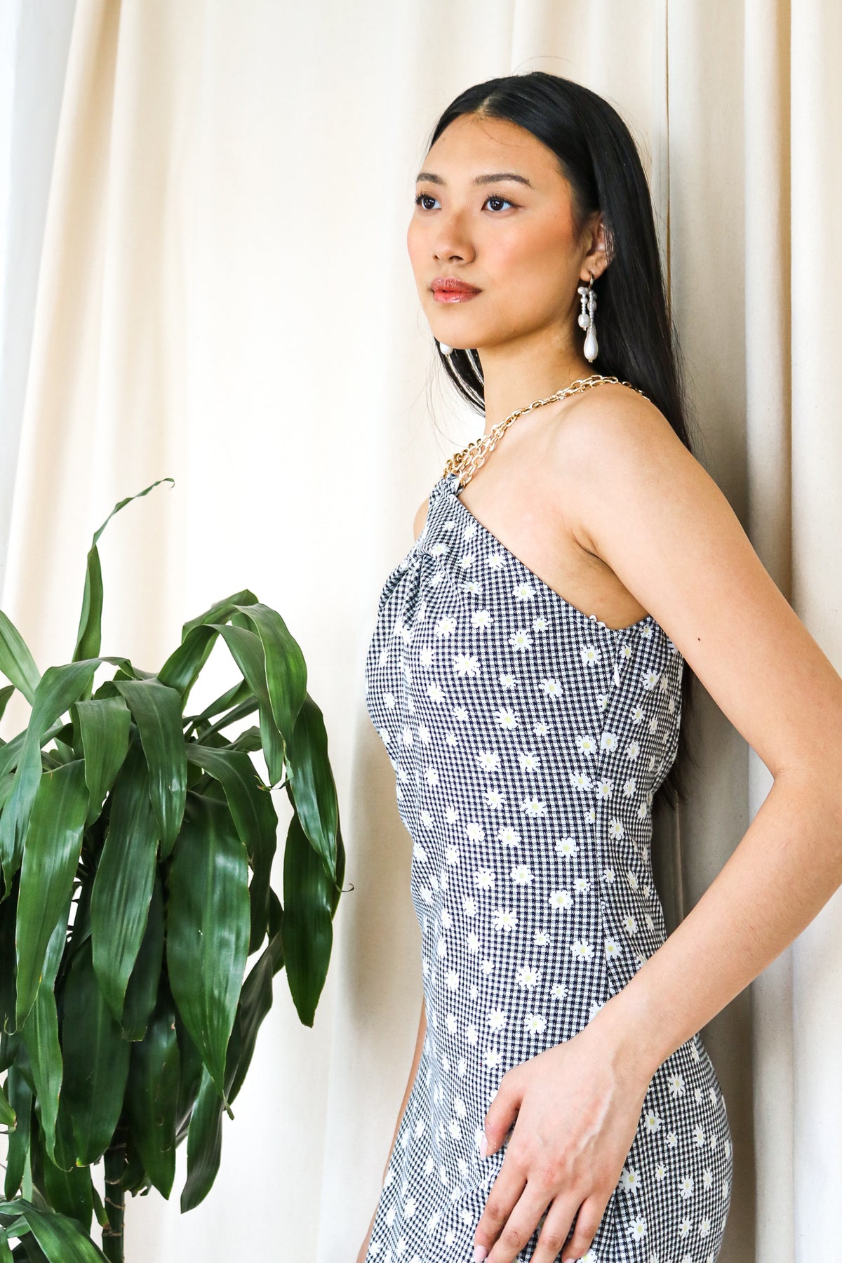 Model is wearing a green gingham midi dress with floral detail and criss cross gold chain detail around the neckline. Left side close up.