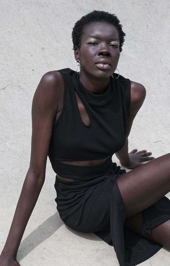 Model wearing a sleek black sleeveless dress with a cut-out detail, seated against a minimalist background.