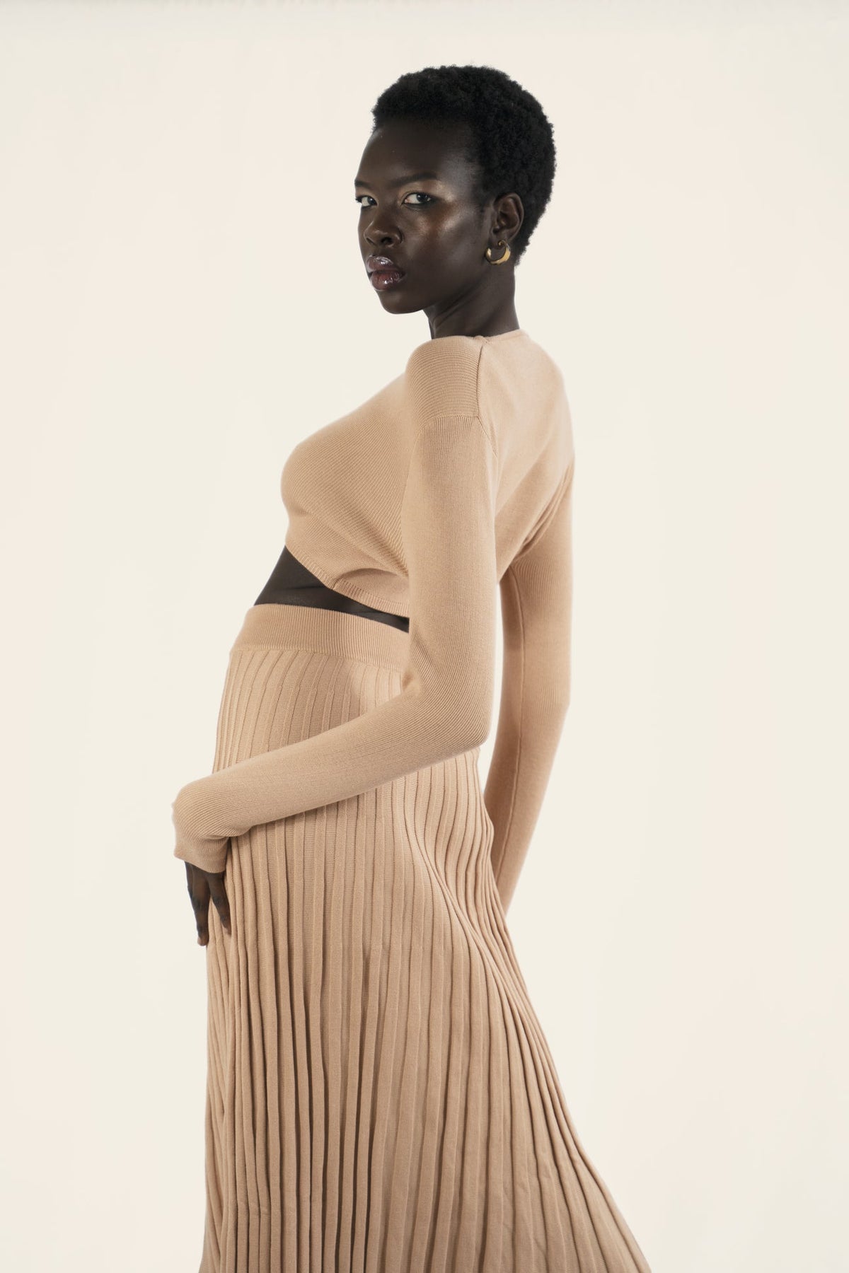 Model wears a nude beige matching set. The ribbed skirt with buttons down the front and a long sleeves plain crop top. Back facing.