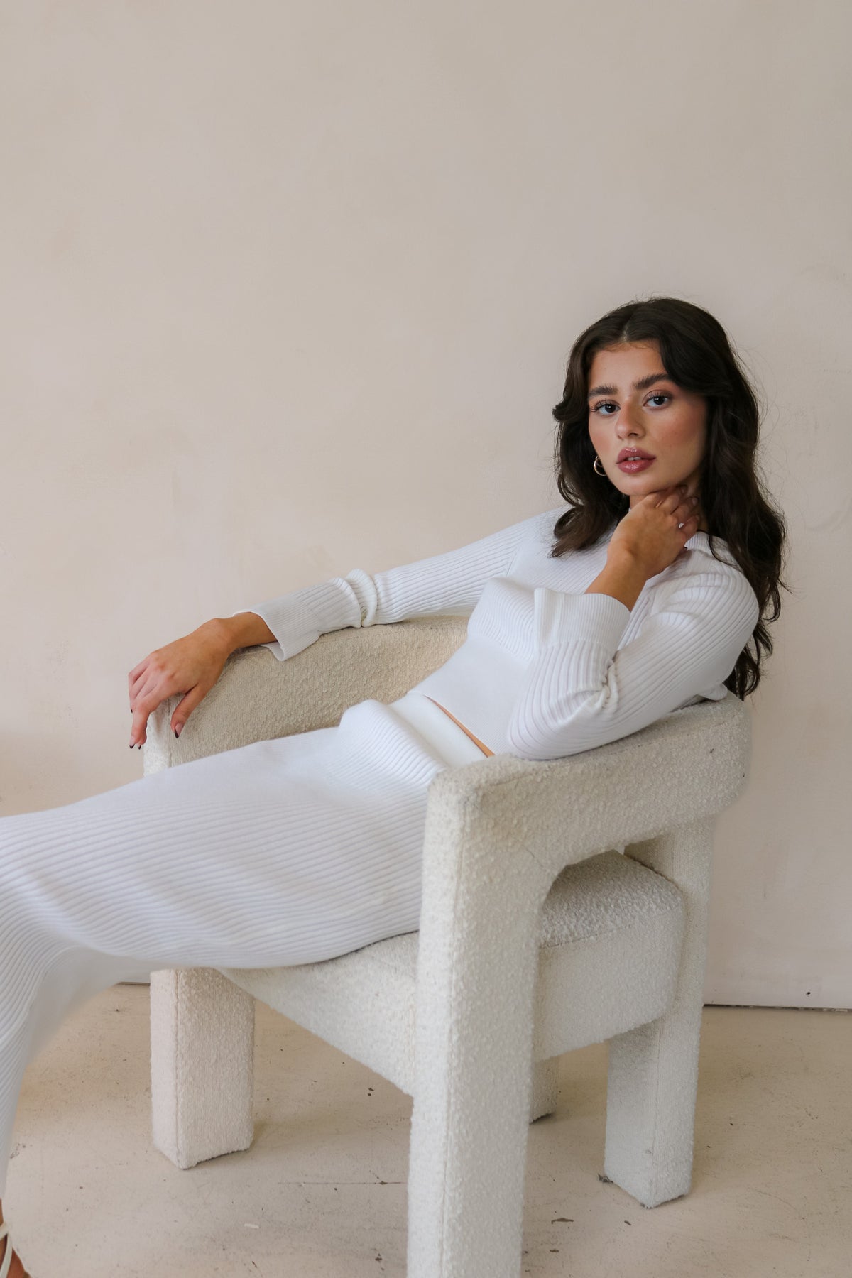 Model is sitting on white chair wearing the matching ribbed sweater and skirt set in white. Front facing.