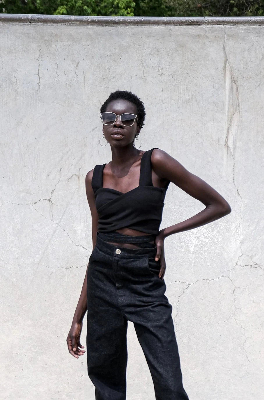 Model stands against minimal background outdoors wearing a sleeveless cross over knitted crop top in black..