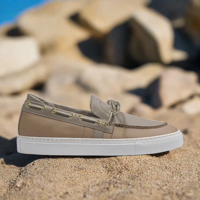 Side view of beige boat loafer sneakers with laces on the side. Outdoors surrounded by sand.  