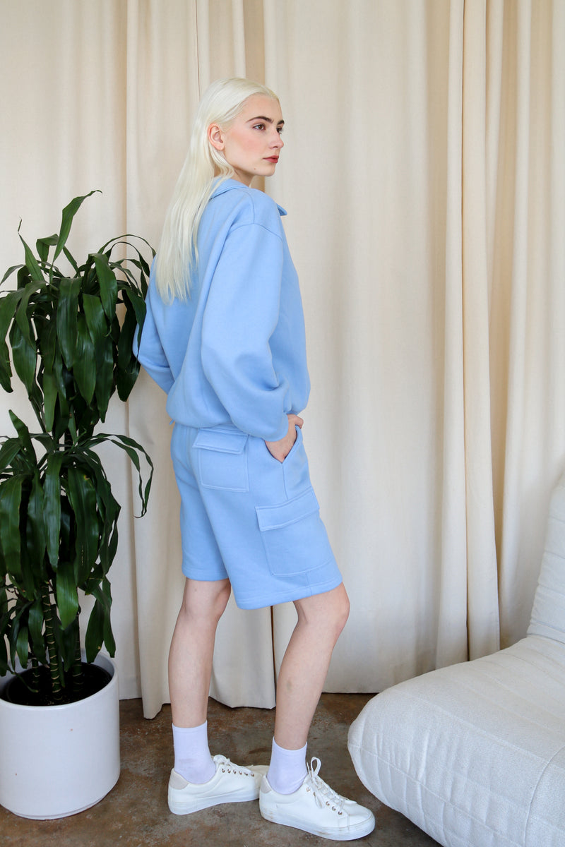 Model wears the blue everyday polo oversized cotton sweatshirt with a polo collar against a neutral backdrop. Side view. 