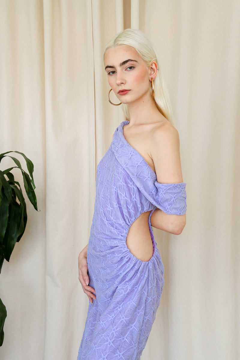 Model wearing a purple one-shoulder textured midi dress, standing beside a plant and a white cushioned chair.