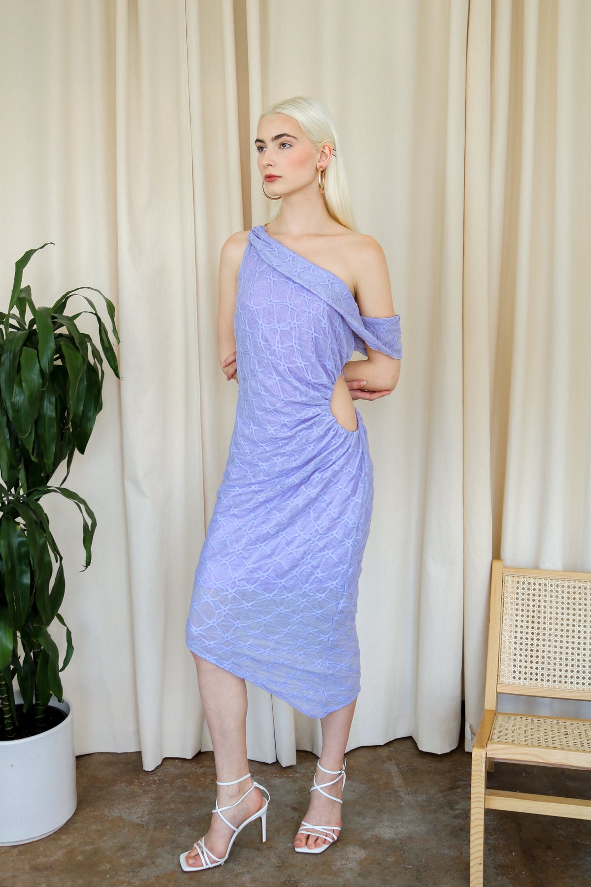 Model wearing a purple one-shoulder textured midi dress, standing beside a plant and a white cushioned chair.