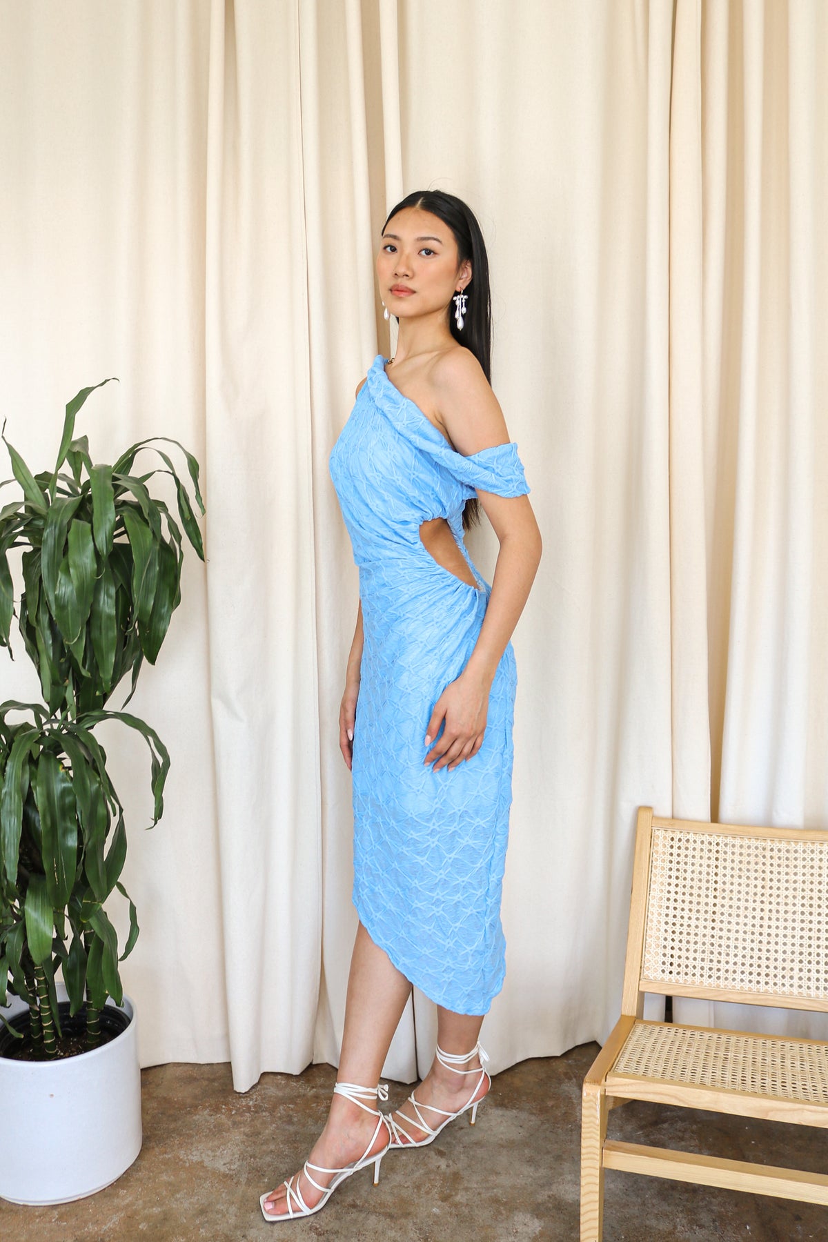 Model wearing a cerulean blue one-shoulder textured midi dress, standing beside a plant and a white cushioned chair.