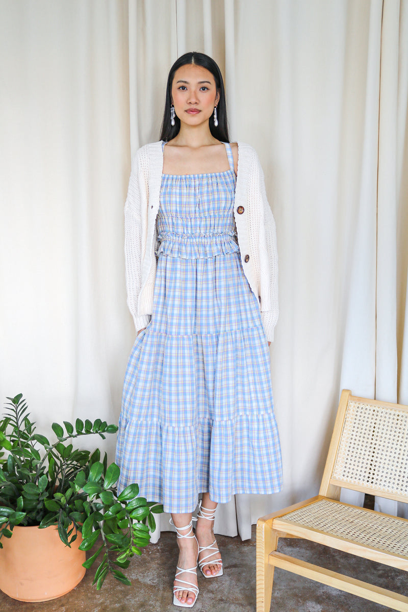 Model wearing a blue plaid smocked tiered maxi dress with a white cardigan, standing beside a plant and a chair with a rattan seat