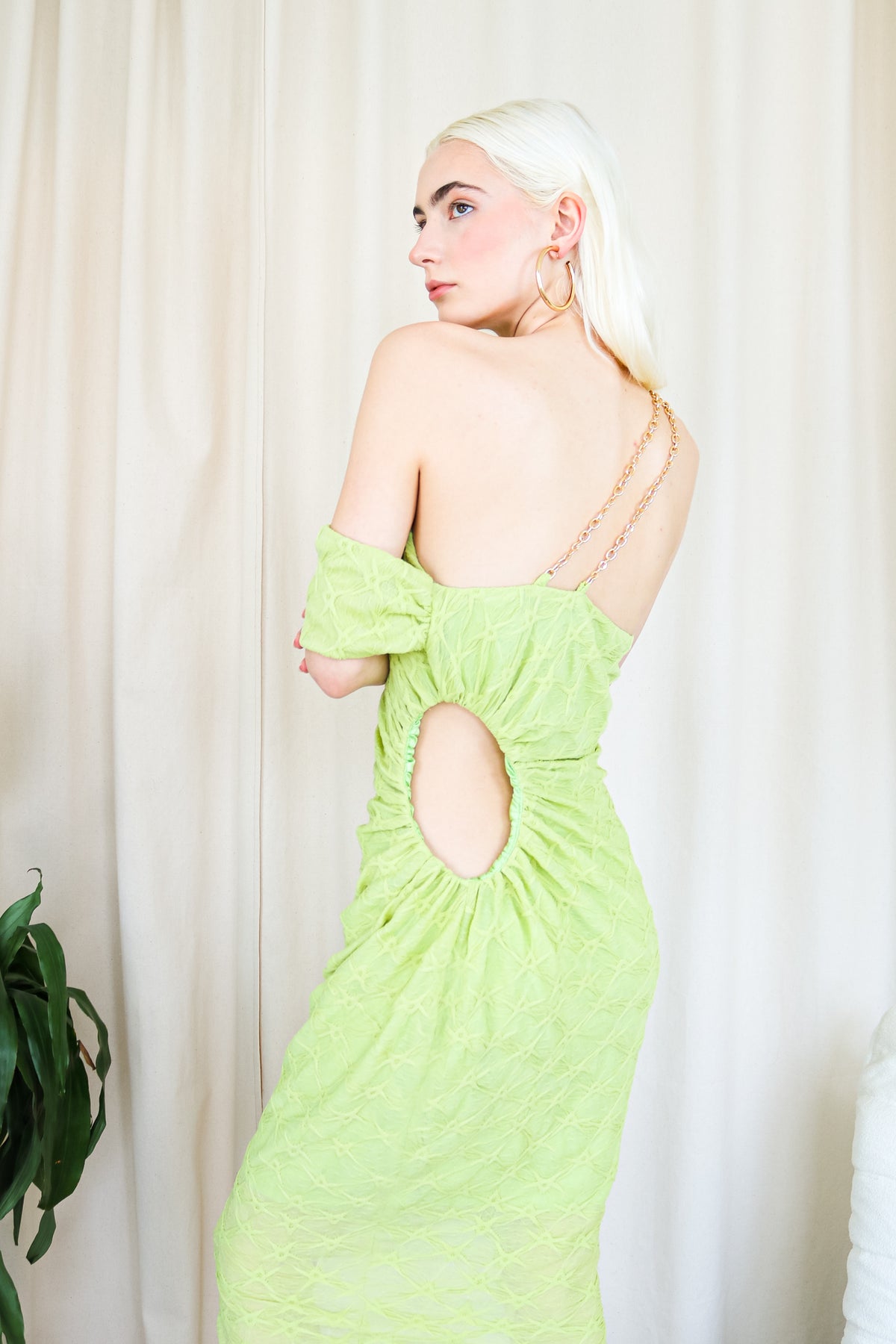 Model wearing a lime green one-shoulder textured midi dress, standing beside a plant and a white cushioned chair. Back.