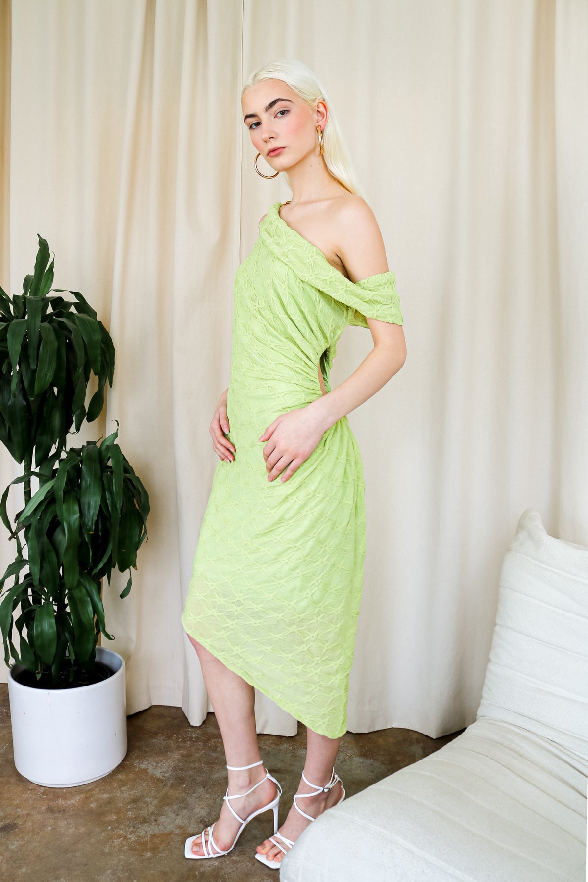 Model wearing a lime green one-shoulder textured midi dress, standing beside a plant and a white cushioned chair.