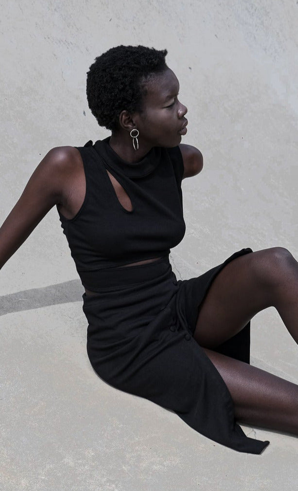 Model wearing a sleek black sleeveless dress with a cut-out detail, seated against a minimalist background.