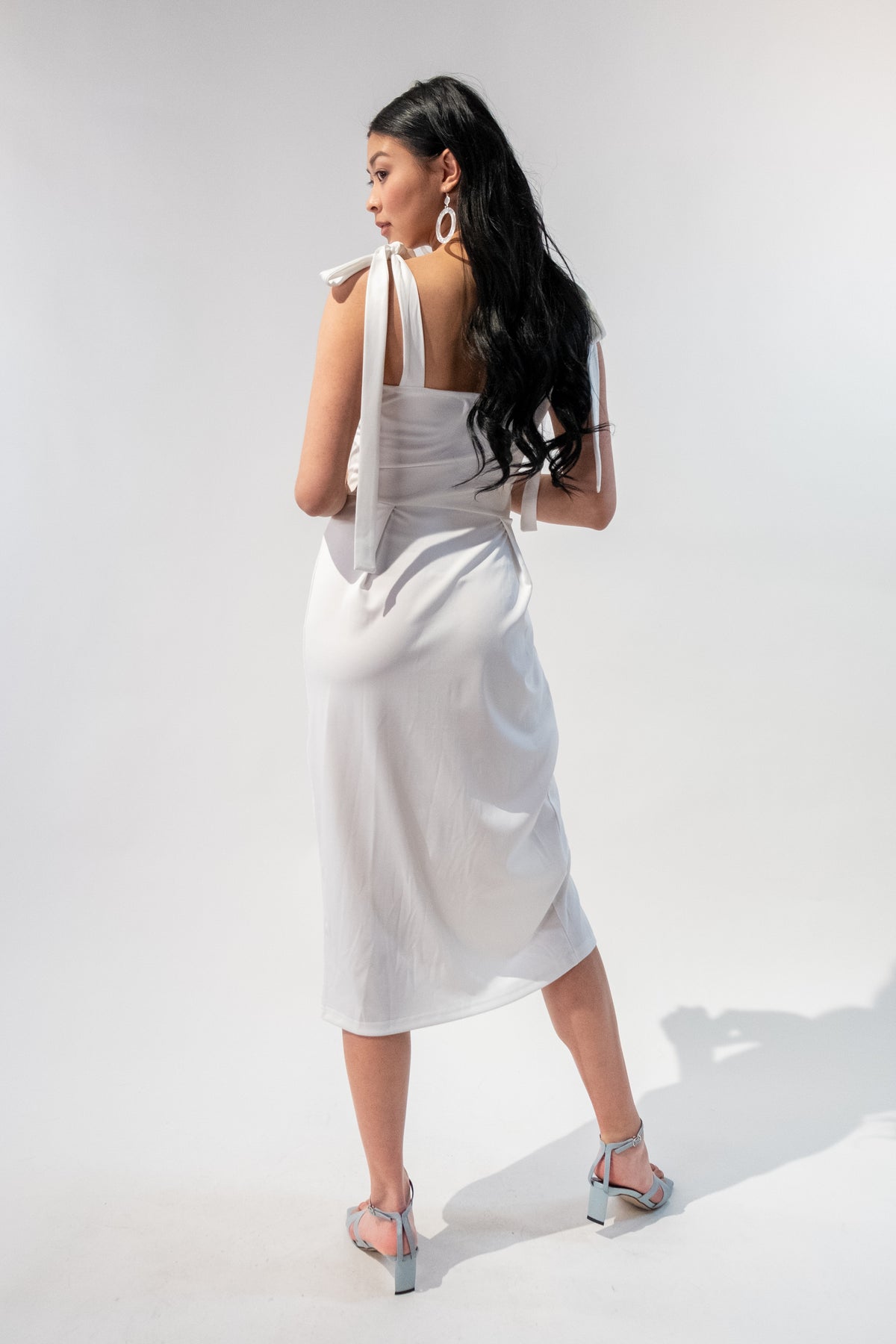 Model standing wearing a tie strap midi dress in white. Back.