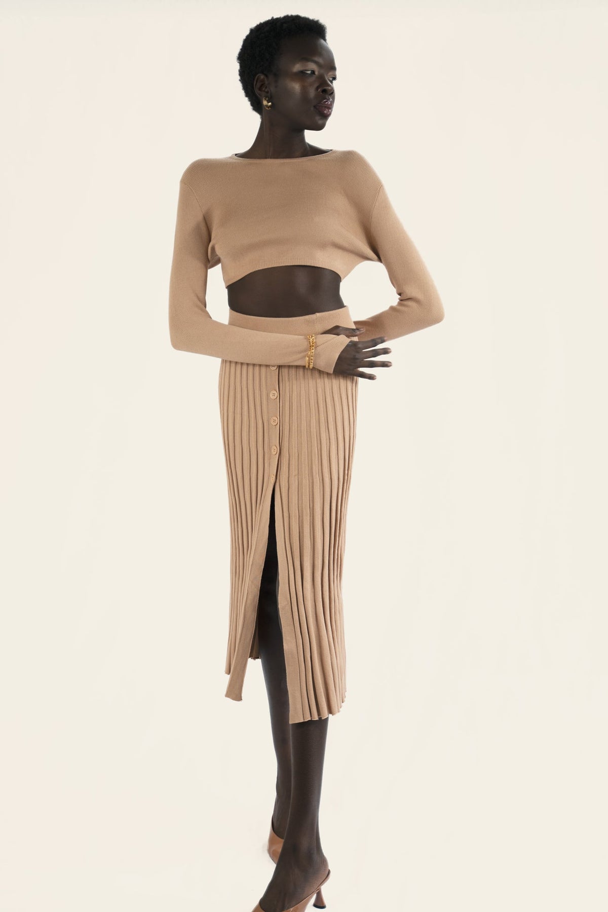 Model wears a nude beige matching set. The ribbed skirt with buttons down the front and a long sleeves plain crop top. Front facing.
