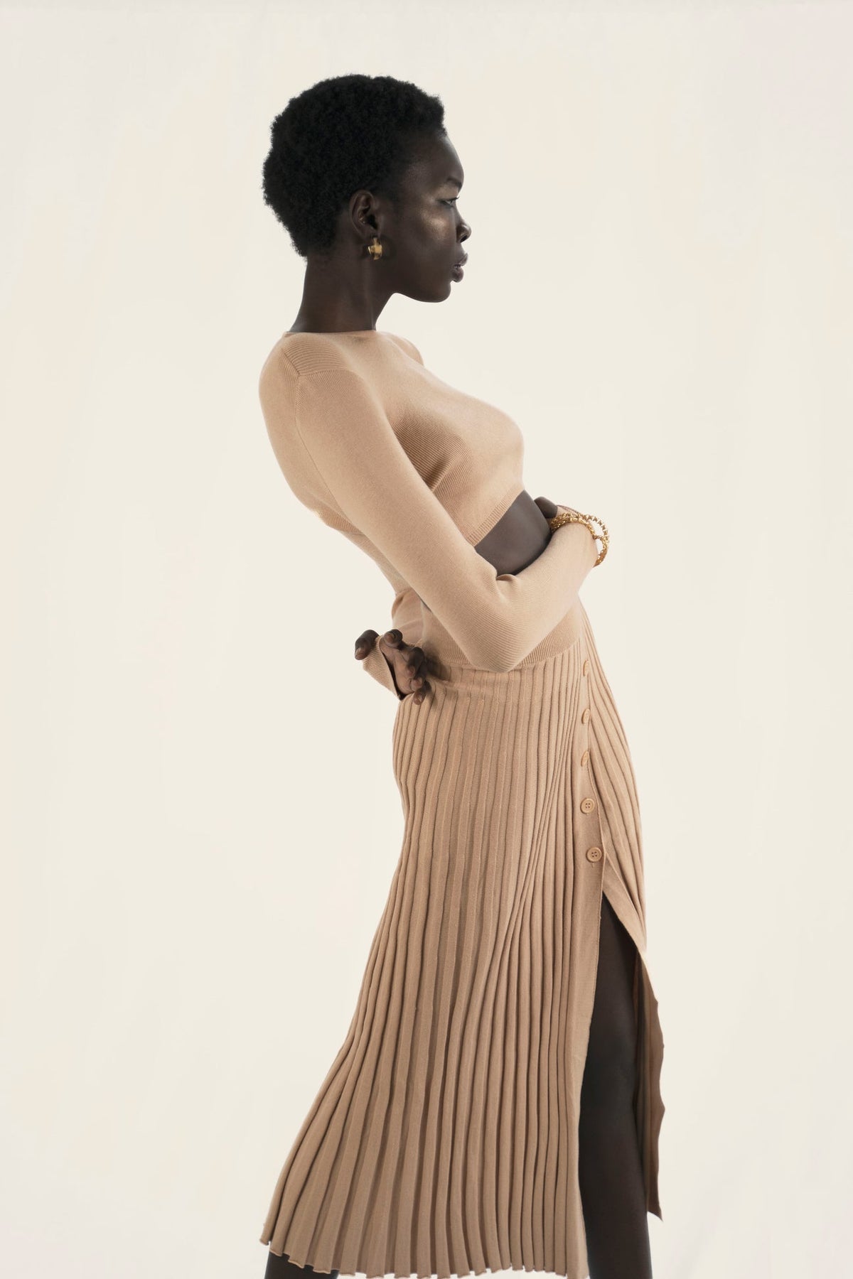 Model wears a nude beige matching set. The ribbed skirt with buttons down the front and a long sleeves plain crop top. Side facing.