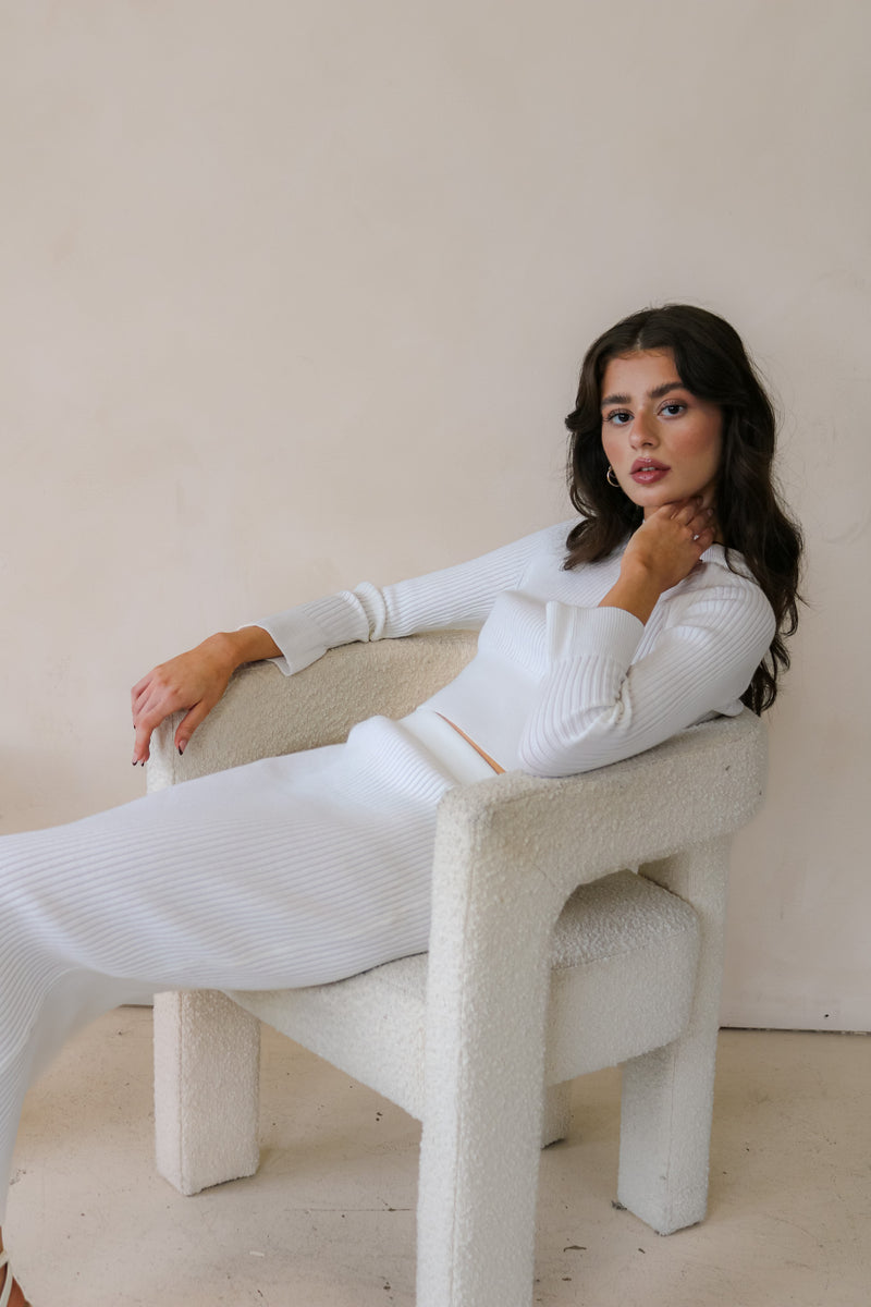 Model is sitting on white chair wearing the matching ribbed sweater and skirt set in white. Front facing. 