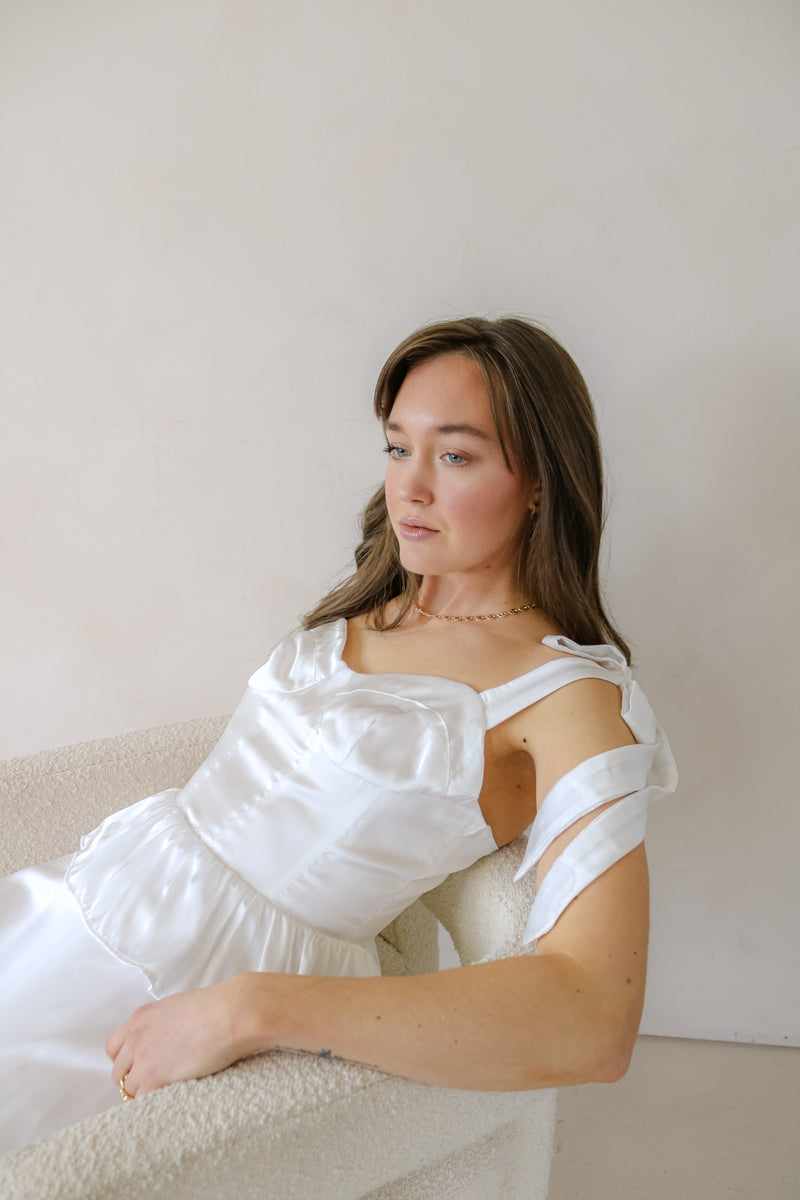 Model wears the Florence Tiered Ruffle Midi dress in white. She is sitting on a chair where you can see the adjustable straps and corset bodice. Front facing.