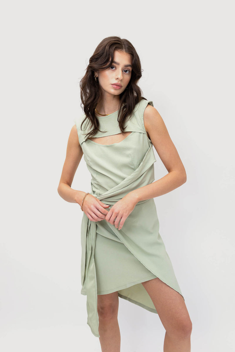 Model wearing an elegant mint green asymmetrical dress with a round neckline, crescent-shaped cutout at the bust, and front tie detail.