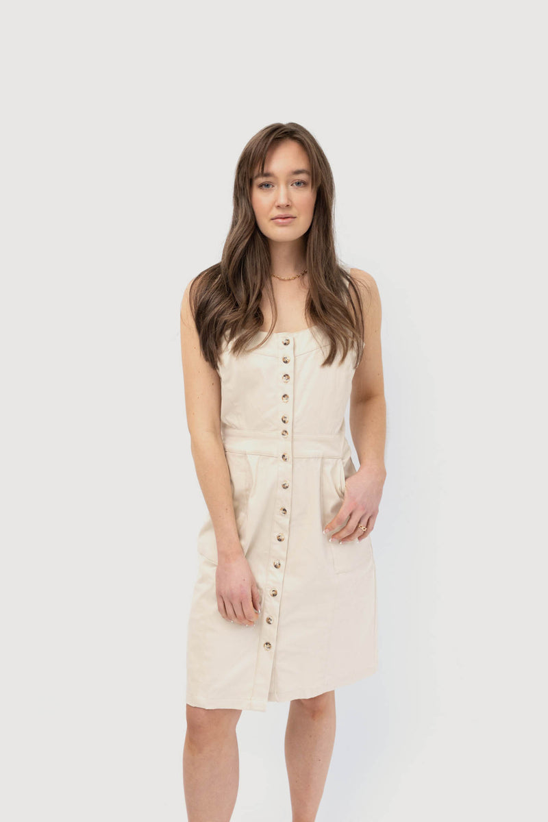 A woman wearing a light beige sleeveless button-down dress with a fitted waist and pockets. The dress features prominent buttons running down the front, giving it a classic and minimalist look. The woman has long brown hair and is standing against a plain white background.