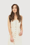 A woman wearing a light beige sleeveless button-down dress with a fitted waist and pockets. The dress features prominent buttons running down the front, giving it a classic and minimalist look. The woman has long brown hair and is standing against a plain white background.