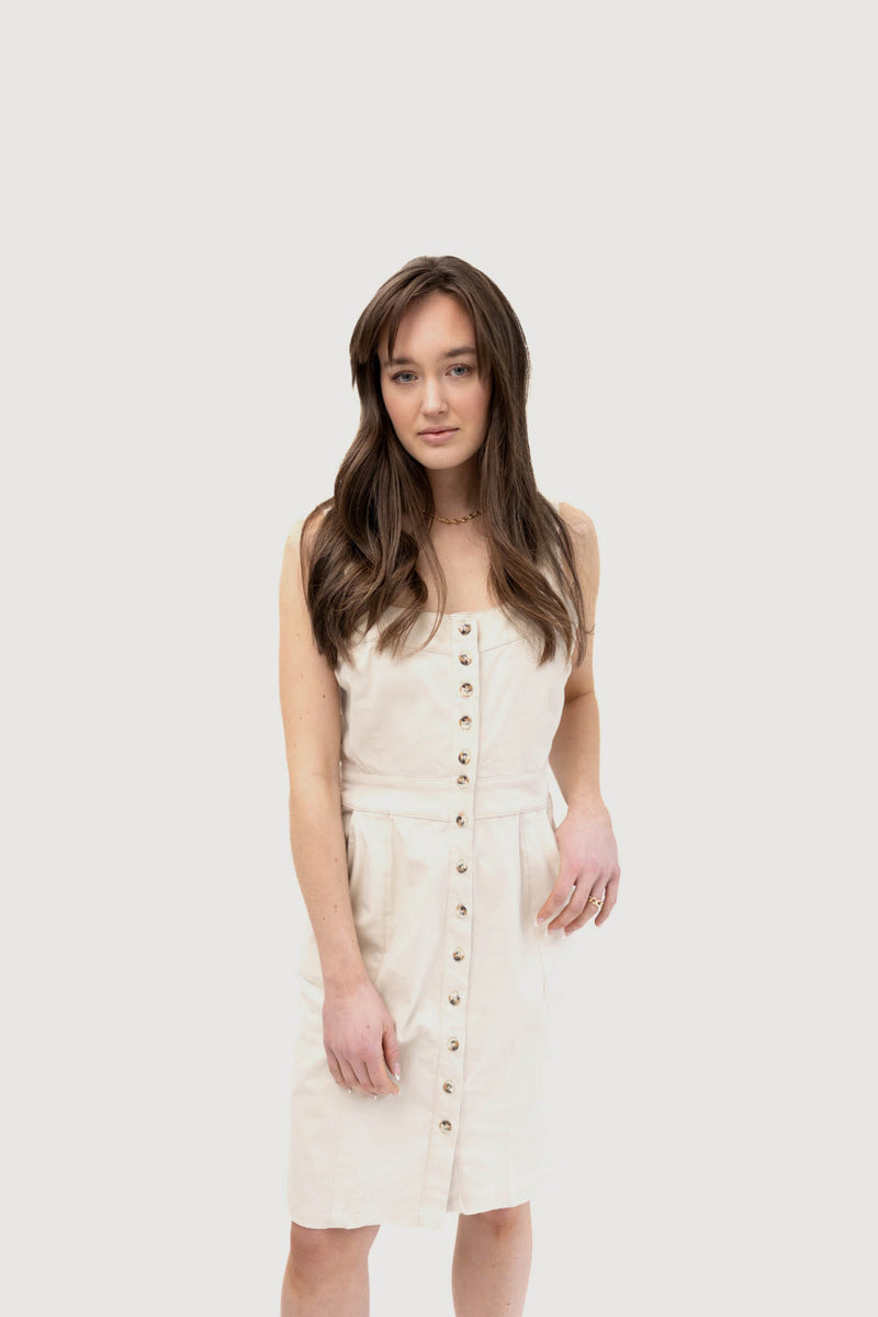 A woman wearing a light beige sleeveless button-down dress with a fitted waist and pockets. The dress features prominent buttons running down the front, giving it a classic and minimalist look. The woman has long brown hair and is standing against a plain white background.