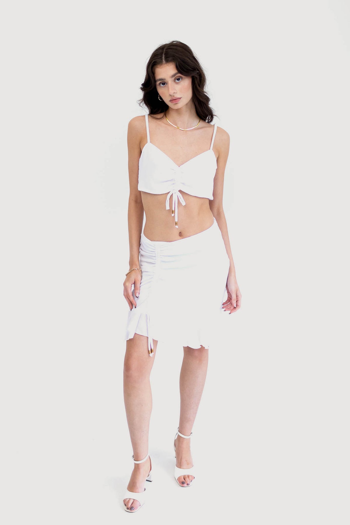 Model wearing a floral ruched crop top with adjustable spaghetti straps and a matching high-waisted skirt with a ruffled hem. White.