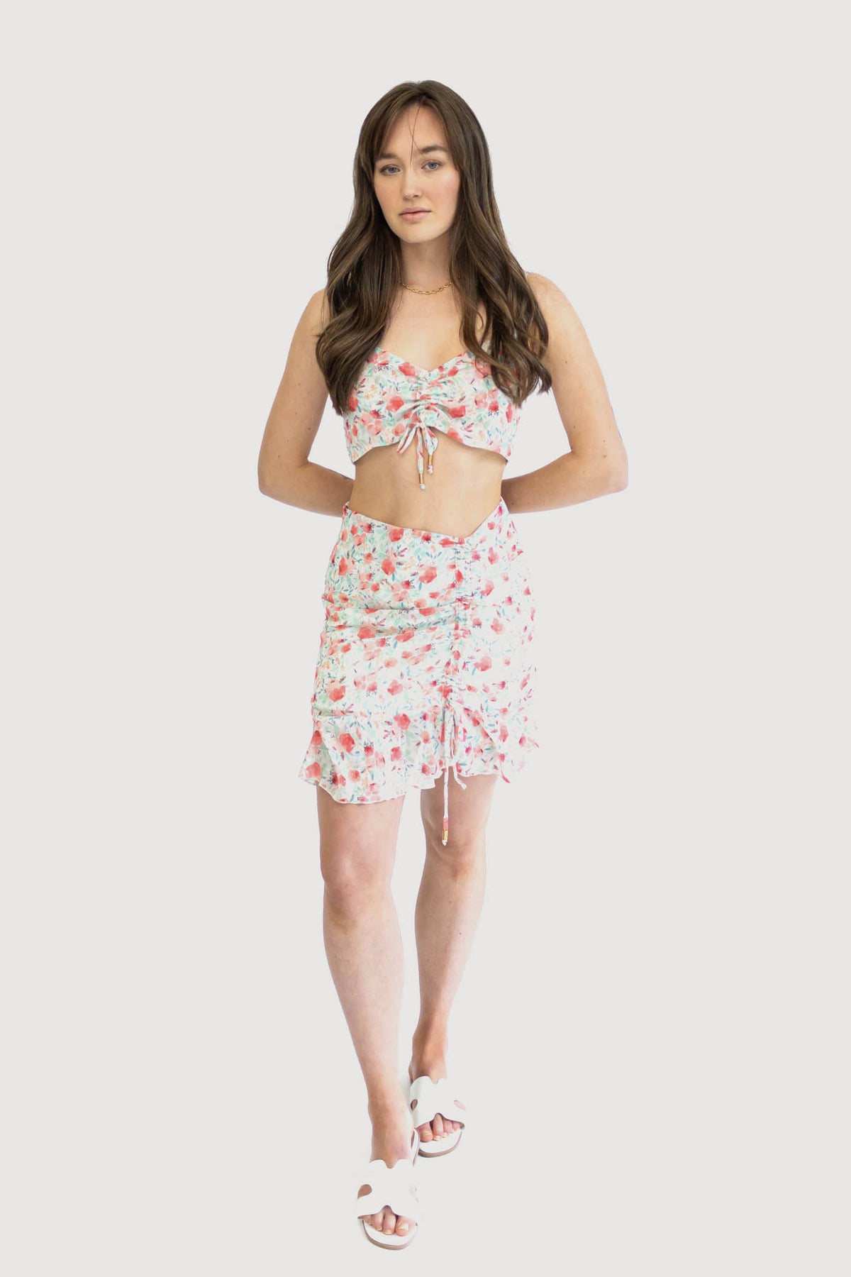 Model wearing a floral ruched crop top with adjustable spaghetti straps and a matching high-waisted skirt with a ruffled hem. Coral peonies.