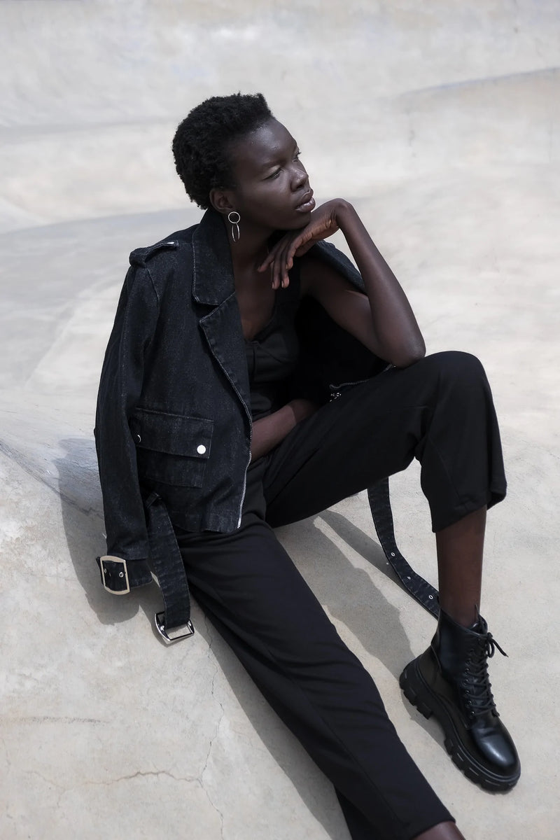 Model wearing an edgy black denim jacket with belted detail, paired with black pants and combat boots, seated in a minimalist setting.